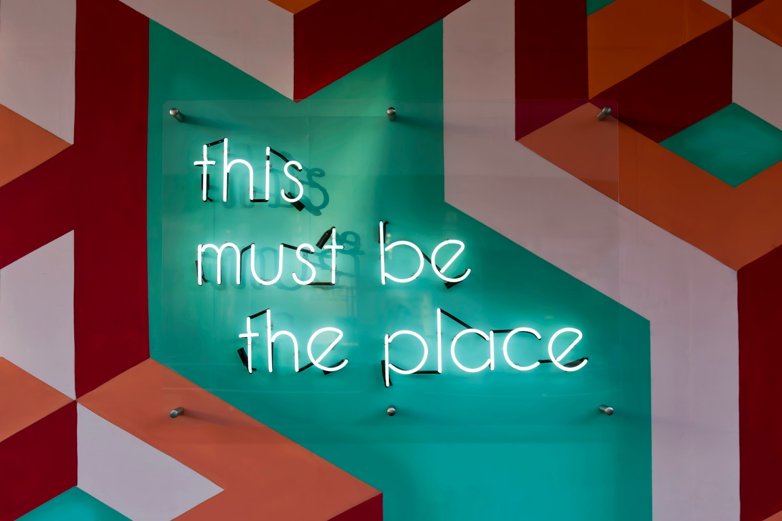 Neon signage that says "This Must Be The Place" on a geometrical background of pink and orange shapes